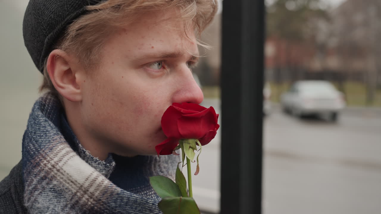 Ung kunstner ved bussholdeplass. En tankefull ung mann står ved en urban bussholdeplass og holder en blomst. Den ettertenksomme kaukasiske kunstneren tar en pause med blomsten mens han venter på bussen midt i byens vinterlandskap.