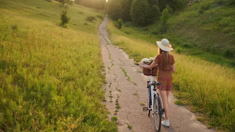 Rückansicht-In-Zeitlupe:-Eine-Schöne-Blondine-In-Einem-Kleid-Mit-Blumen-In-Einem-Korb-Und-Einem-Retro-Fahrrad-Geht-Auf-Der-Sommerwiese-Die-Straße-Entlang,-Schaut-Sich-Um-Und-Lächelt-Mit-Einem-Gefühl-Der-Freiheit.