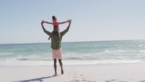 padre afroamericano sonriente llevando a su hija en una playa soleada