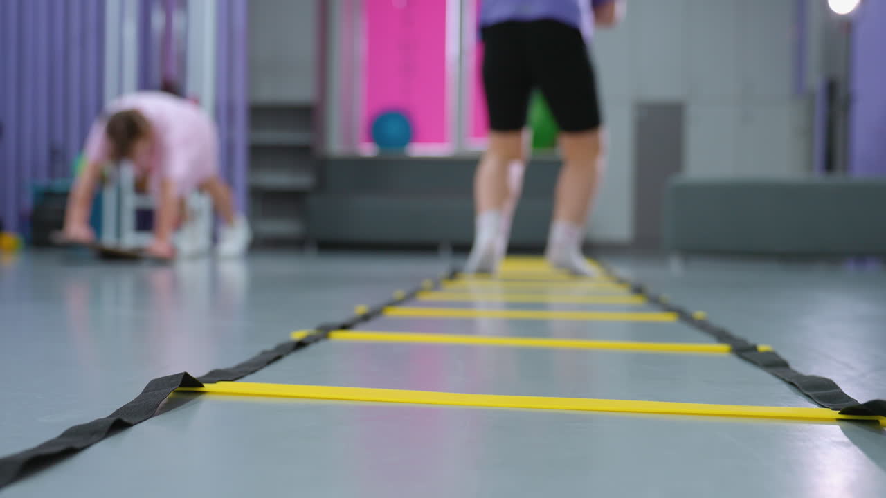 vista trasera de la dama de púrpura saltando escalera de agilidad para la velocidad y la coordinación mientras otro niño en el fondo borroso trabaja en el equilibrio, varios equipos de gimnasio y herramientas de entrenamiento visibles