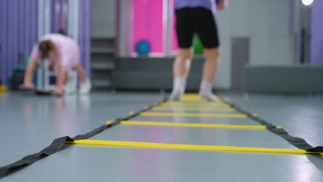 vista trasera de la dama de púrpura saltando escalera de agilidad para la velocidad y la coordinación mientras otro niño en el fondo borroso trabaja en el equilibrio, varios equipos de gimnasio y herramientas de entrenamiento visibles