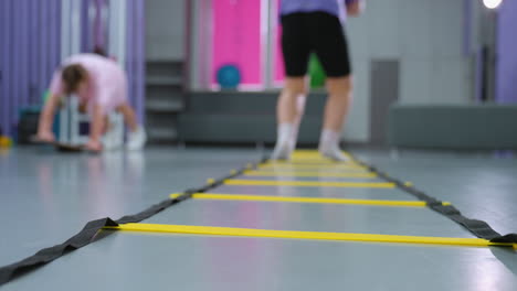 back view of lady in purple jumping agility ladder for speed and coordination while another kid in blurred background works on balance, various gym equipment and training tools visible