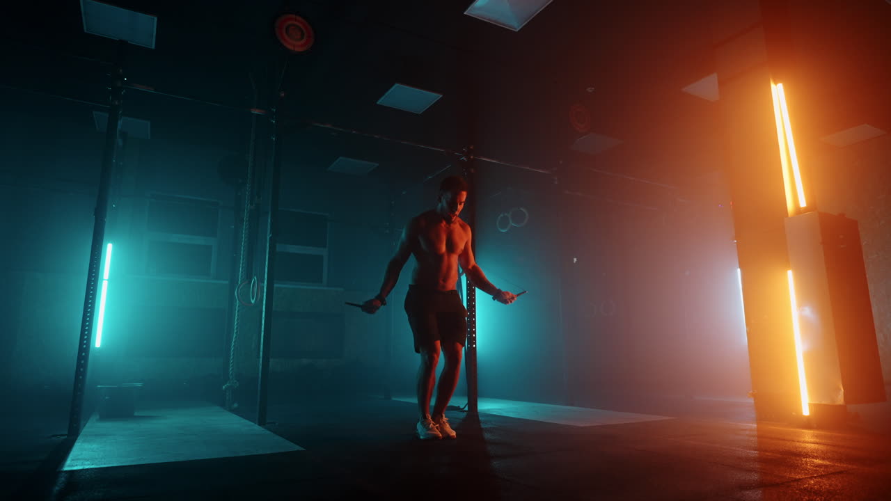 Muscular man with bare torso is jumping using a skipping rope. Male boxer with skipping rope. Fighter doing jump rope exercises in colorful light with smoke in slow motion.