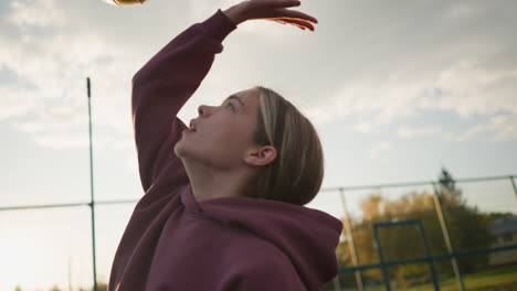 woman in hoodie throws volleyball up to serve in outdoor court with open field in background, person walks in distance while sunlight creates warm atmosphere