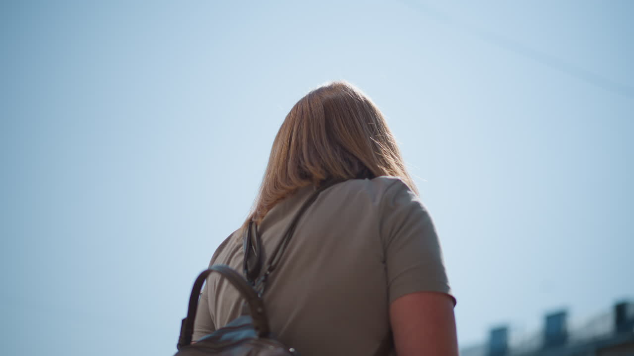Lower view of solitary observer standing under bright sunlight near school building, looking around, showing sadness and reflection during quiet summer day outdoors in city