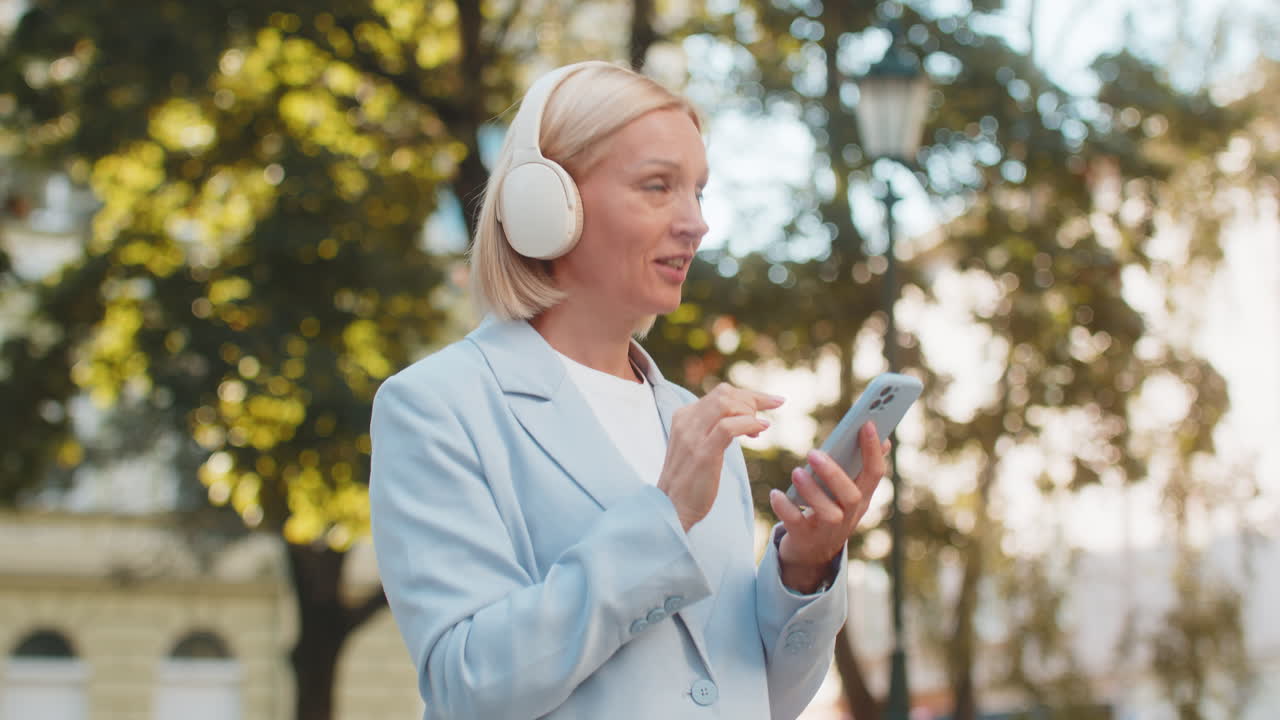 Happy smiling mature businesswoman with smartphone listening to music in headphones on city street