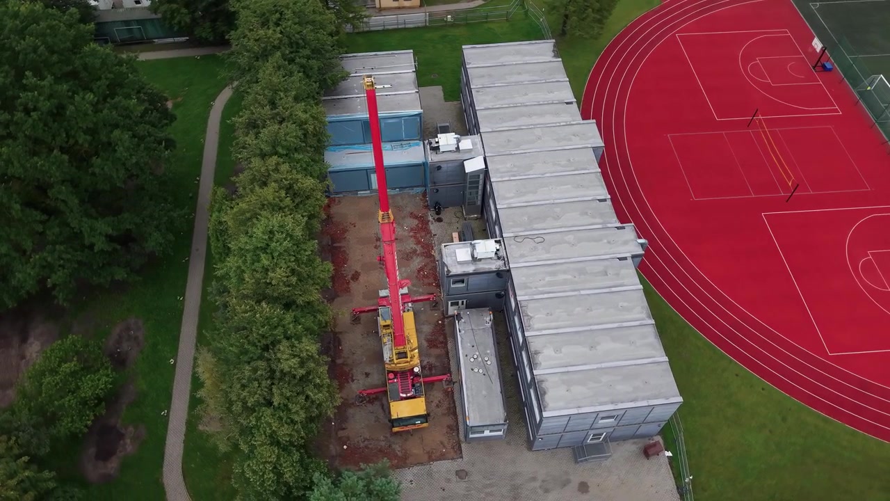 Luftoptagelser viser en byggekran, der samler modulbygninger ved siden af en skoles sportsplads og løbebane, omgivet af frodige grønne træer på en overskyet dag.
