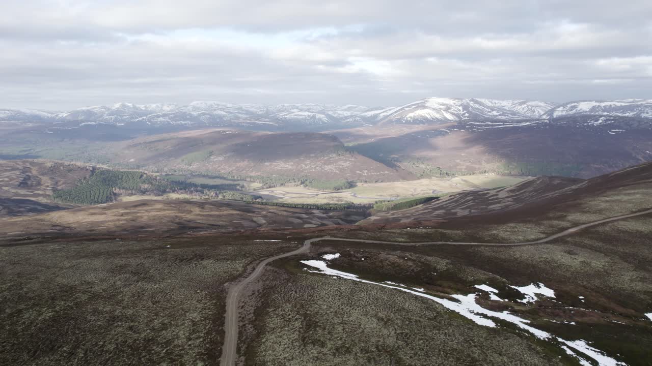 drone aéreo cinematográfico que desciende hacia una pista 4x4 y páramos de brezo y brezo de liquen mirando hacia un paisaje montañoso con parches de nieve y nubes