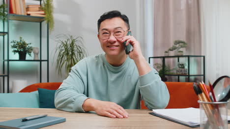 happy excited asian man making phone conversation call with friends sitting on couch at home room