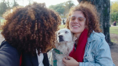 two joyous young women sitting with dog in park and video calling