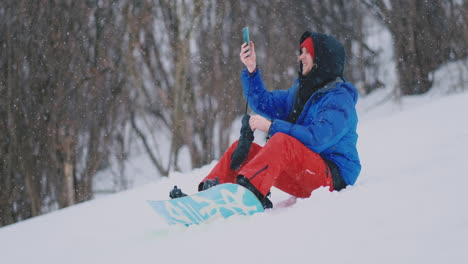 male snowboarder sitting on the snow taking photos on the phone beautiful scenery of the resort for social networks