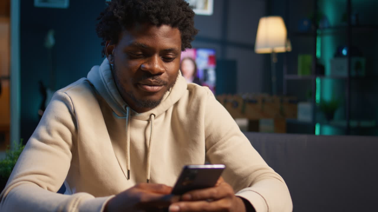 African American man on a sofa texting mates in his living room, entertained by social media and messages. Depiction of remote work breaks, digital communication and relaxation at home. Camera B.