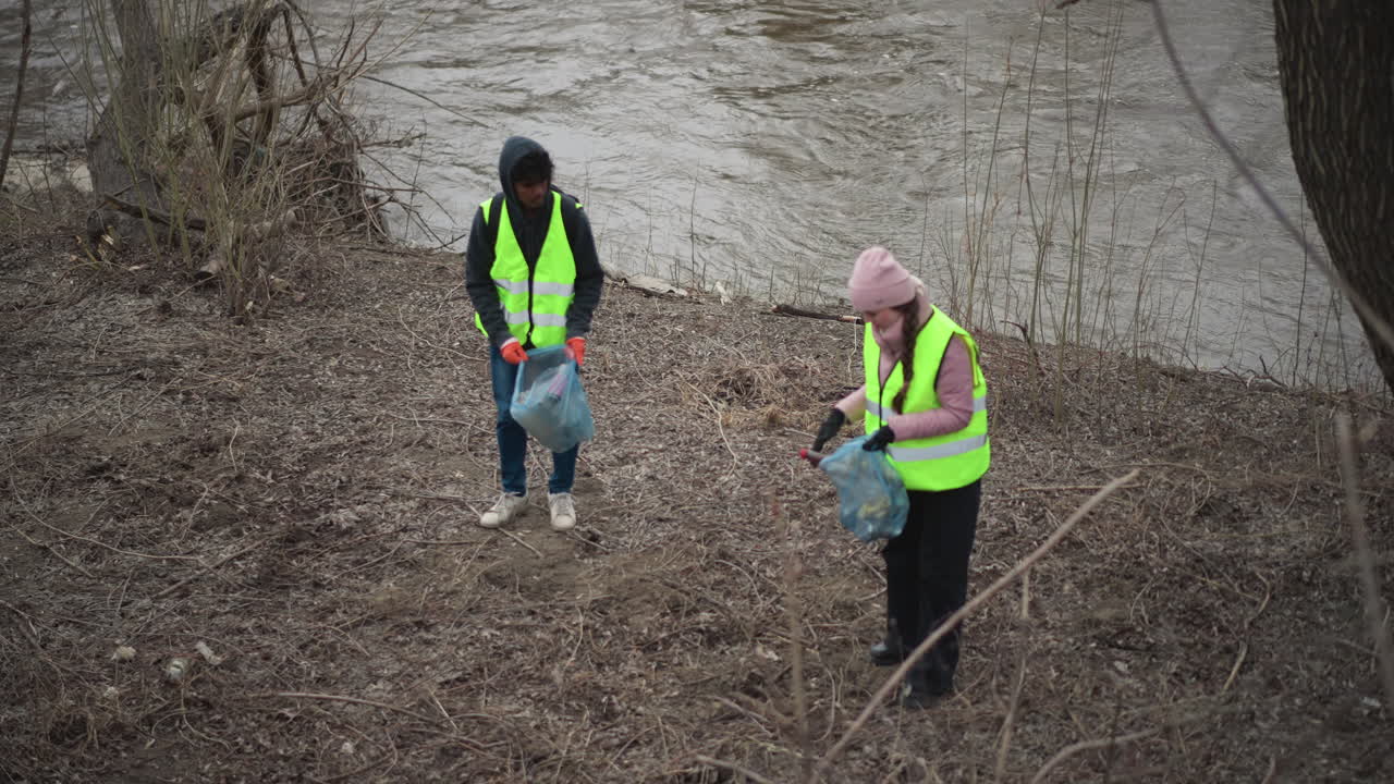 Volunteers in reflective vests collecting plastic bottles and trash along polluted riverbank during environmental cleanup on cold overcast day, wearing gloves and carrying bags to protect nature