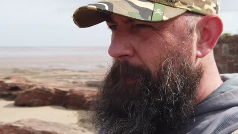 hombre caucásico barbudo con gorra de béisbol sintiendo ansiedad mirando a través de la playa ventosa