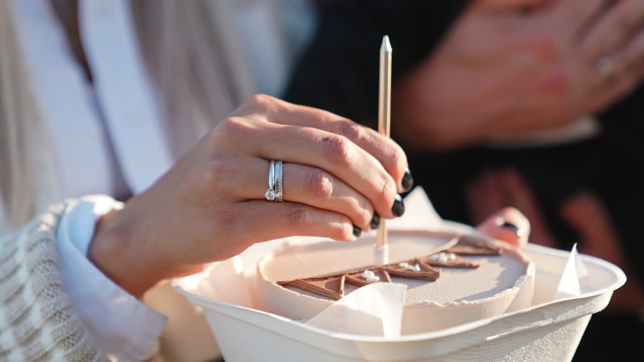 Female hand puts long single candle into the cake. Close up. Unrecognized male holding a baby at backdrop.