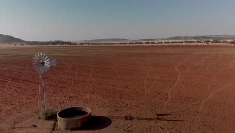 Aerial-dolly-shot-of-a-working-windpump-in-red-dry-field-with-water-in-dam