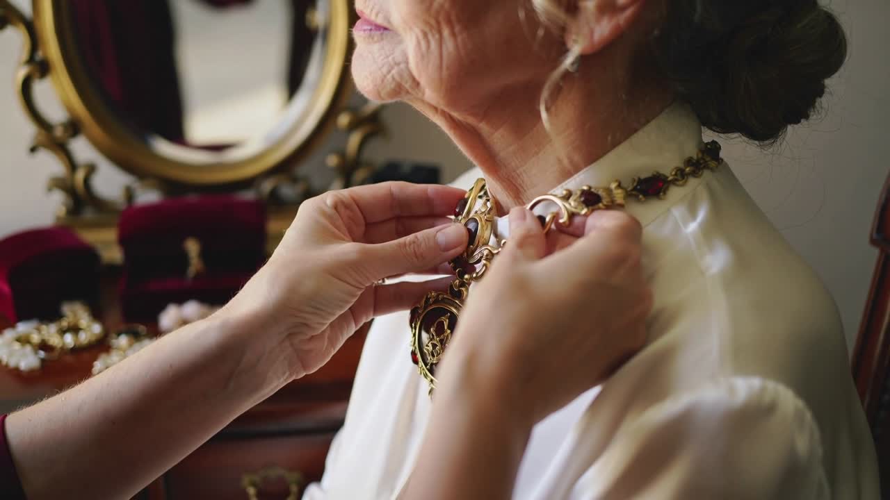 Un assistente che prende una collana d'oro dalla specchiera, assicurandone la chiusura, mentre prepara un'anziana con un top di seta per un evento.