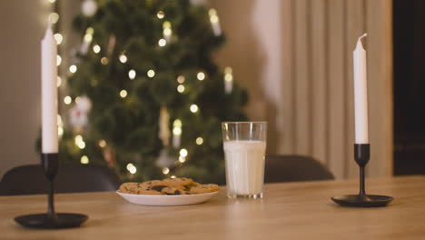 Camera-Focuses-On-An-Glass-Of-Milk-And-A-Plate-Full-Of-Cookies-On-An-Empty-Table-With-Two-Candles-In-A-Room-Decorated-With-A-Christmas-Tree-1