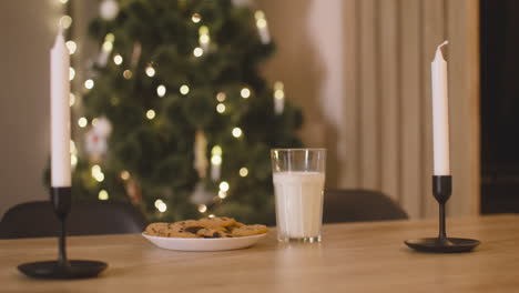 la cámara se enfoca en un vaso de leche y un plato lleno de galletas en una mesa vacía con dos velas en una habitación decorada con un árbol de navidad 1