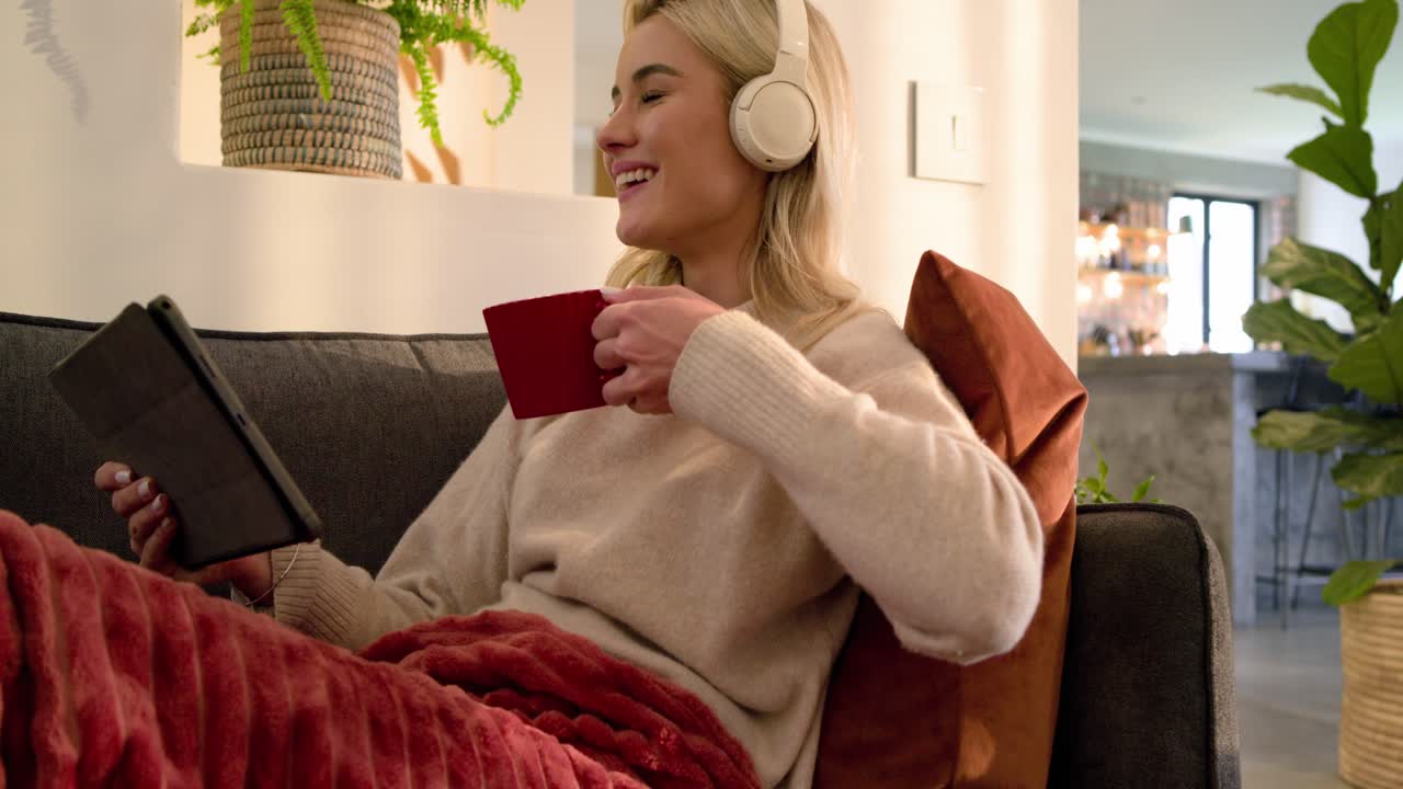 Woman receiving alert scrolling tablet on dark gray couch wearing headphones, laughing, sipping mug