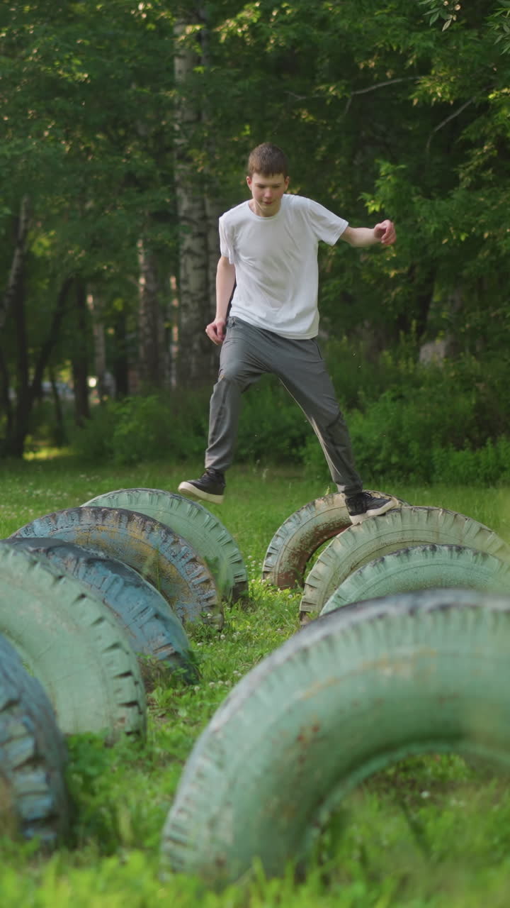 un niño en una camisa blanca y pantalones grises corre a través de las dos primeras filas de neumáticos en un campo cubierto de hierba, capturado a cámara lenta mientras se detiene en el último neumático en la fila central