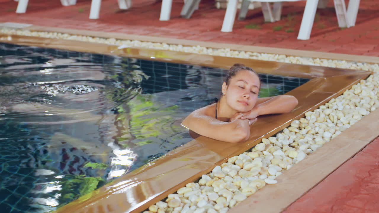 Donna che si rilassa al bordo della piscina dell'isola, viaggiatrice che appoggia il mento sulle braccia, lievi increspature dell'acqua, vasca della piscina piastrellata, bordo di ciottoli, sedie a sdraio rosse, superficie riflettente, bagliore serale, atmosfera di vacanza tranquilla.