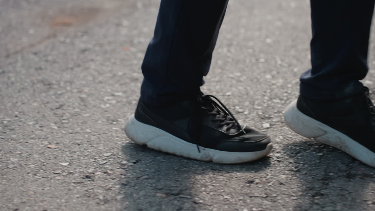 sneakers walking on cracked pavement, man in dark jeans taking slow commuter stroll, closeup of shoe soles and laces, morning light casting long shadow, textured asphalt and small weeds at curb