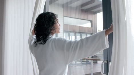 Mujer-Birracial-Usando-Albornoz-Abriendo-Cortinas-Y-Mirando-Por-La-Ventana-En-Casa,-Cámara-Lenta