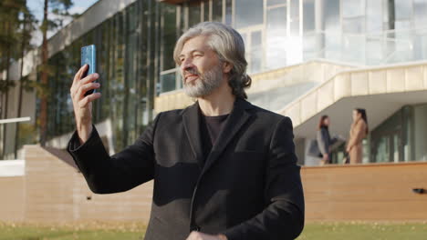 smiling mature man waves during a video call on his smartphone outdoors