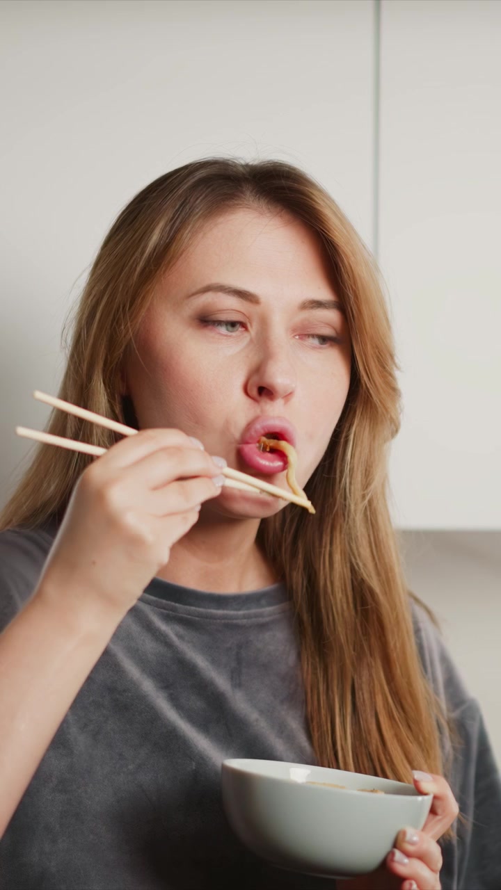 Close up of woman using chopsticks to eat noodles from bowl. scene is set in indoor kitchen with soft lighting focusing on action and texture detail of eating. Background details include