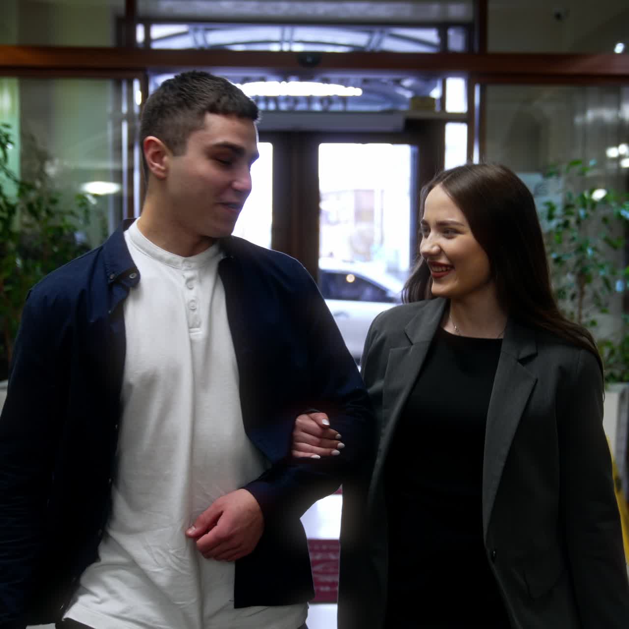 A Caucasian couple walks in through the sliding door of a hotel. Smiling people go along the lobby talking and looking at each other