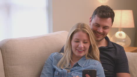 Evening-shot-of-couple-relaxing-on-sofa-at-home-looking-at-mobile-phone---shot-in-slow-motion