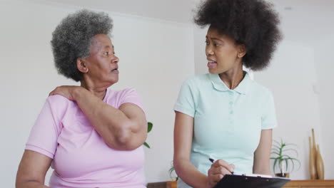 African-american-female-physiotherapist-wearing-face-mask-helping-senior-female-patient-exercise