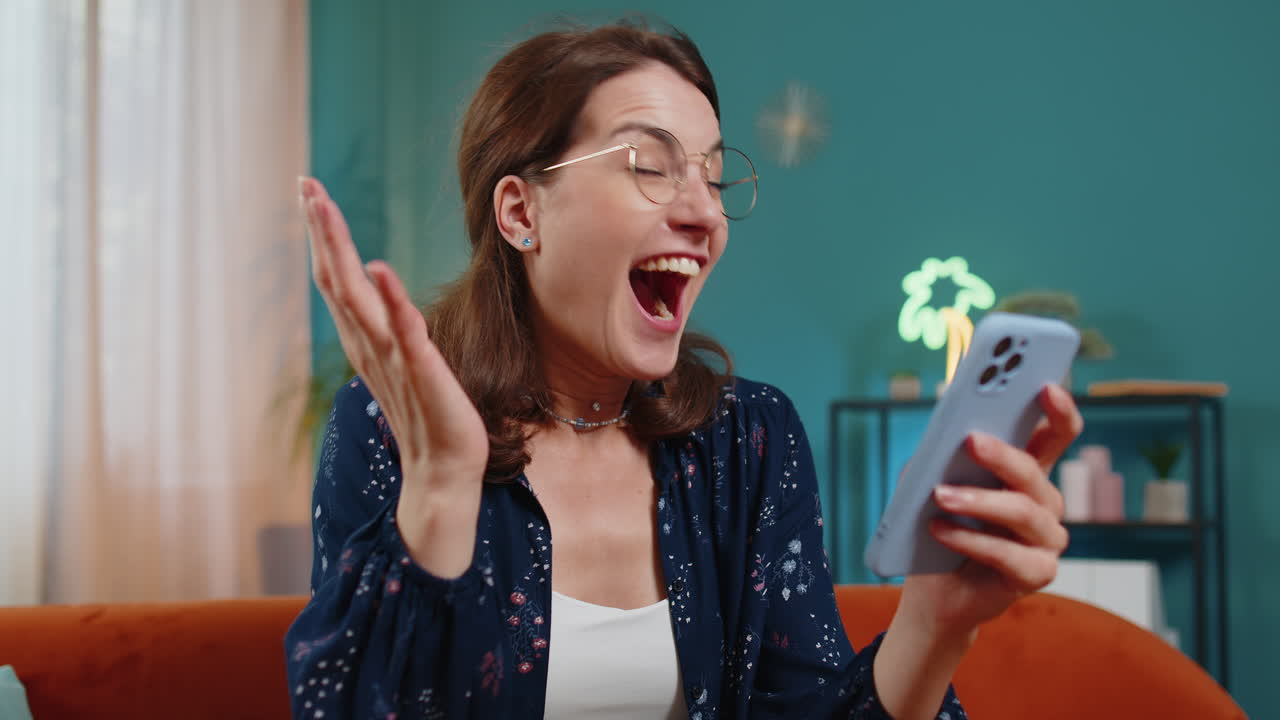 Excited happy amazed winner woman using smartphone receive good news celebrate victory at home