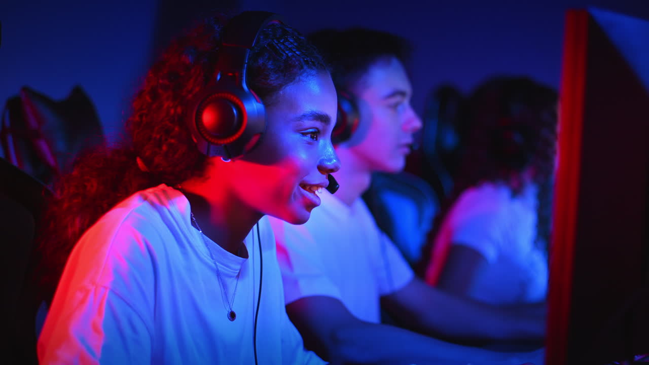 White boy and black girl teens in headsets playing video games in video game club with blue and red illumination, talking in voice chat. Slow motion