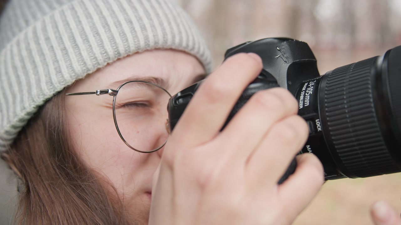 Lähikuva naisvalokuvaajasta rajaamassa DSLR-kameralla ulkona, pipo ja silmälasit päässä, vakaat kädet säätämässä objektiivia, pehmeä talvinen tausta, lämpimät hillityt sävyt, keskittynyt ilme, aito dokumentaarinen tyyli