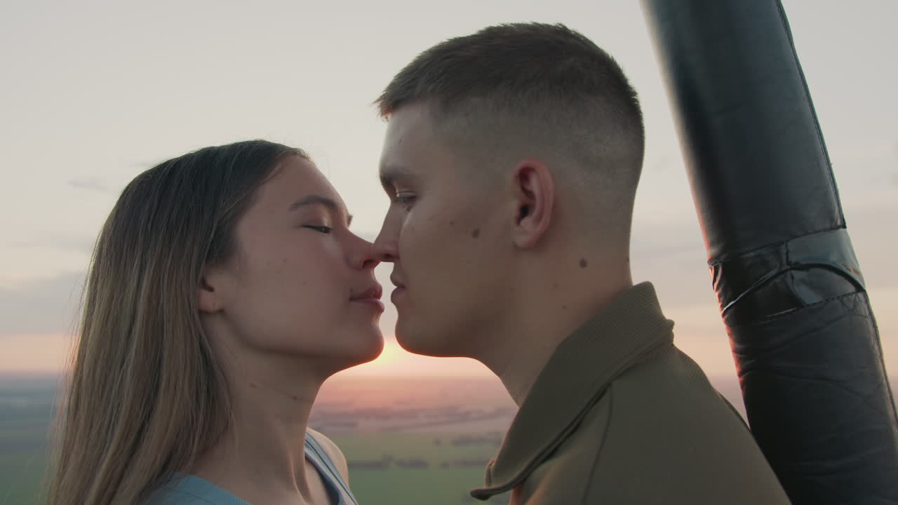 close up couple in hot air balloon embracing and resting head on shoulder while floating above patchwork fields at golden sunset with soft sky hues capturing romantic adventure moment together