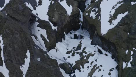 Volando-Cerca-De-La-Ladera-De-Una-Montaña-Cubierta-De-Nieve-Para-Revelar-Una-Pequeña-Cascada-Que-Arroja-Nieve-Derretida-En-La-Montaña