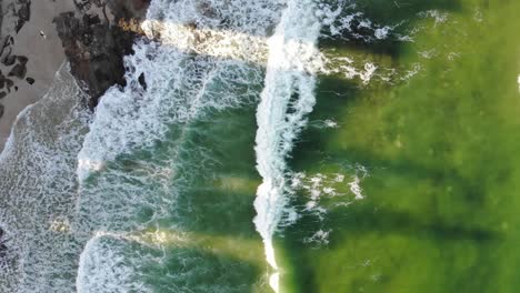 a lime green ocean mixed with hints of dark blue pushes small waves into the dark rocks sat between the pale, coarse sand whilst shadows cast from apartment buildings block the sunsets afternoon glow