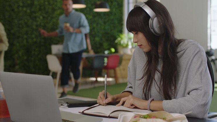 giovane donna d'affari asiatica che scrive appunti godendo dello studio ascoltando musica indossando cuffie studentessa che lavora in un ufficio multietnico affollato
