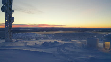 reverse aerial view through snow covered cabin and tower on wintry scandinavian hillside summit at sunrise