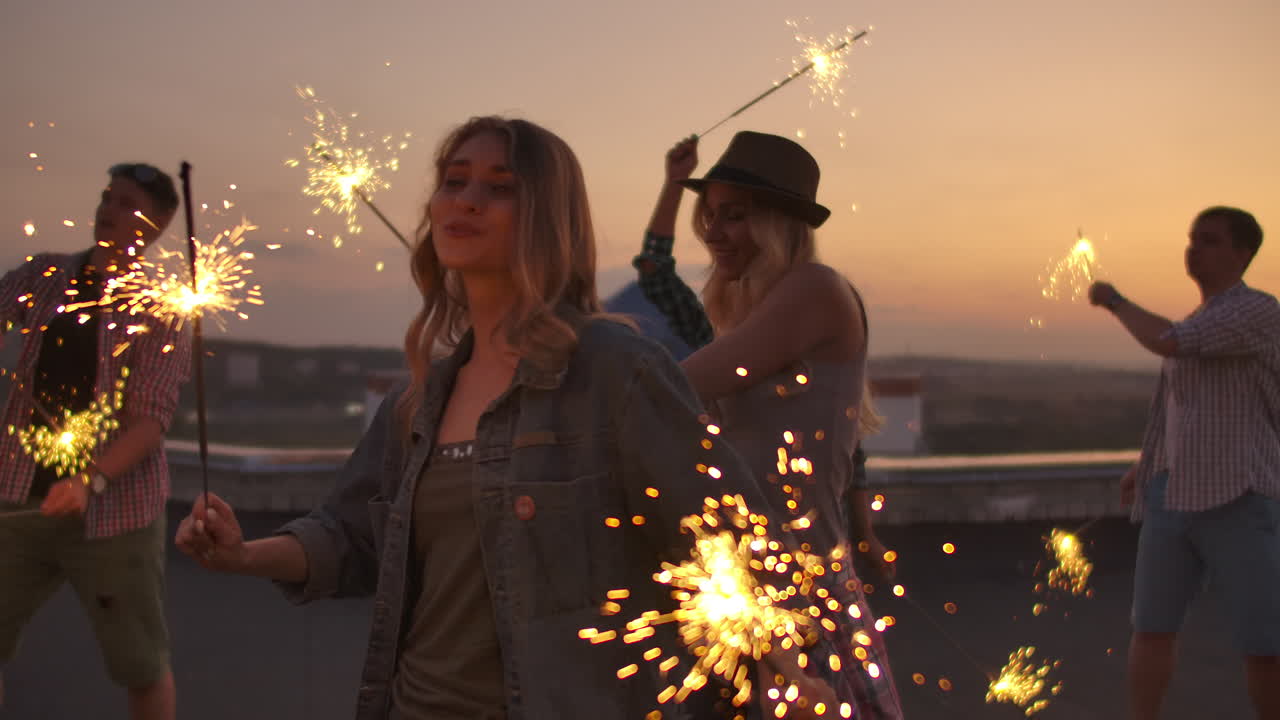 Female on the roof moves her arms and body beautifully and dance with her friends on a summer evening with big bengal light. Her hair blows beautifully in the wind.