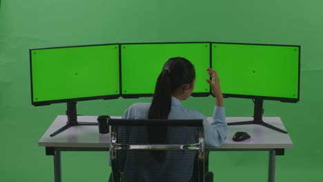 녹색 스크린 스튜디오에서 모의 여러 컴퓨터 모니터와 함께 작업하는 동안 두통을 가진 아시아 여성의 뒷면