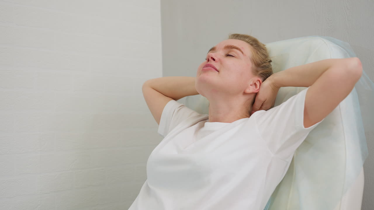 Client with hair tied in bun reclines on salon chair, gently raising hands in relaxed manner while supporting head, set against soft blur interior wall creating calm self-care atmosphere