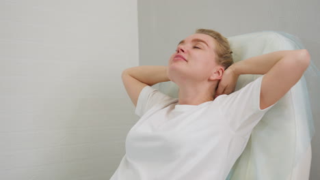 client with hair tied in bun reclines on salon chair, gently raising hands in relaxed manner while supporting head, set against soft blur interior wall creating calm self-care atmosphere