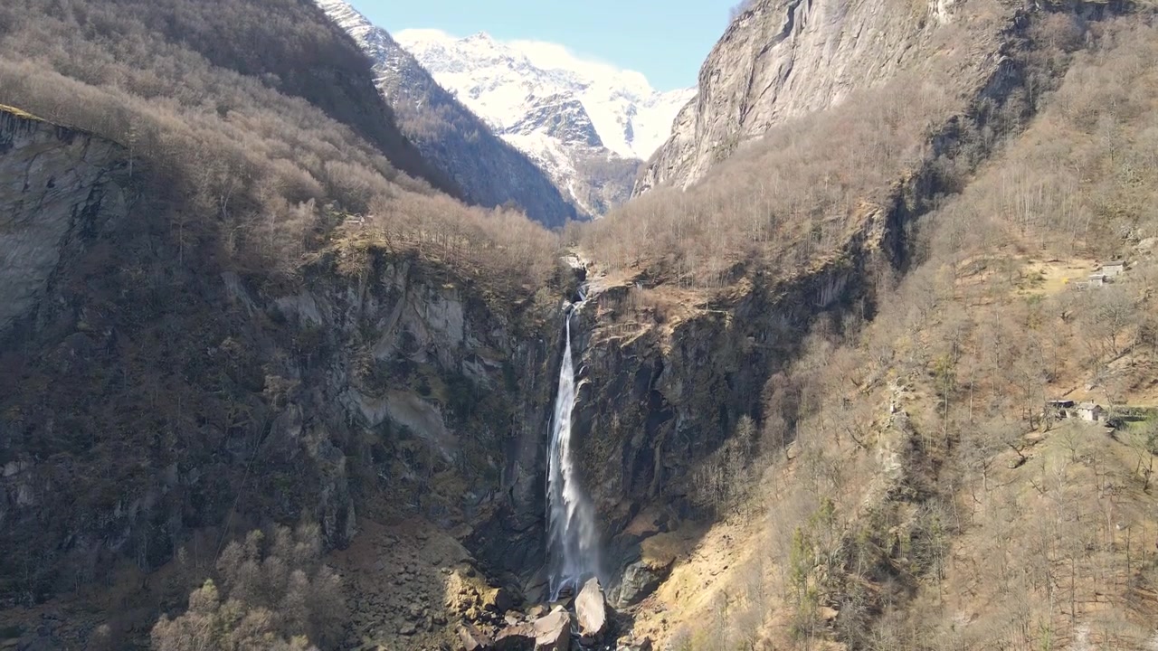 Cascata di Foroglio che precipita da un ripido dirupo in una remota valle svizzera, circondata da montagne innevate e alberi spogli, e il piccolo villaggio, Foroglio, Ticino, Svizzera, inquadratura dal basso