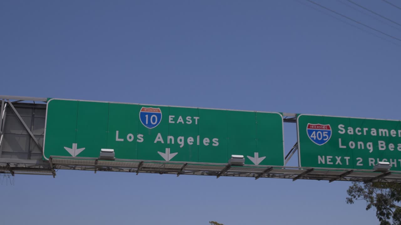 Close-up of Los Angeles freeway interchange sign with directions to Los Angeles, Sacramento, and Long Beach. The bright blue sky serves as the backdrop for the highway signage
