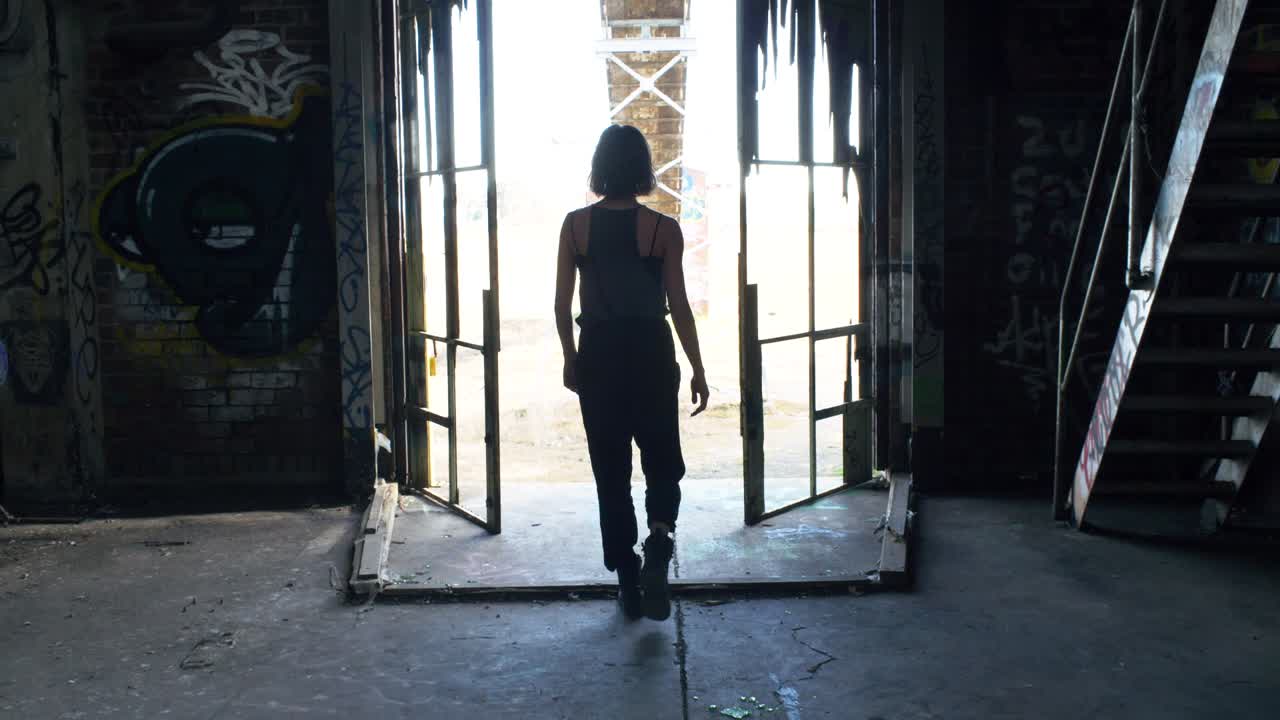 Woman In Grunge Fashion Clothing Walking Towards Exit Of A Rundown Warehouse - medium shot
