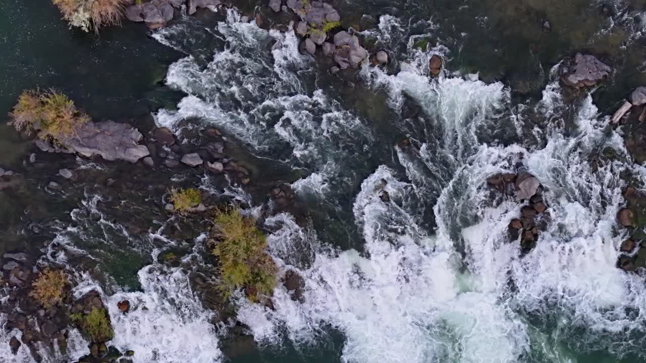 Aerial close-up of the Snake River's rapids in Idaho, capturing churning whitewater cascading over rocks and boulders in a narrow channel, with dark green water swirling around the rugged landscape