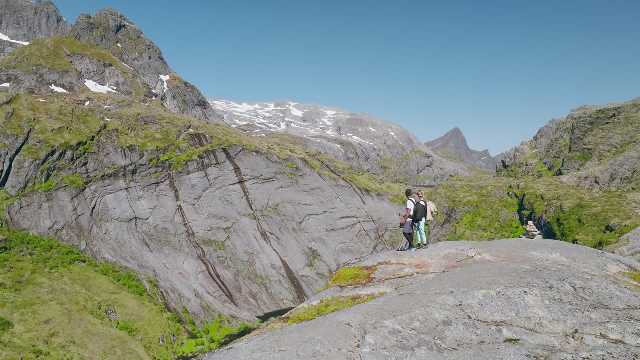 Una coppia fa escursioni su aspre rocce di granito con montagne innevate in lontananza sotto un cielo azzurro limpido, catturando avventura e bellezza paesaggistica in Norvegia.